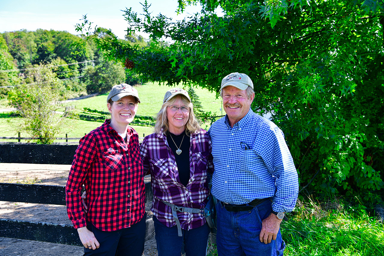 Three people standing outside in front of a lush field of green grass and trees.