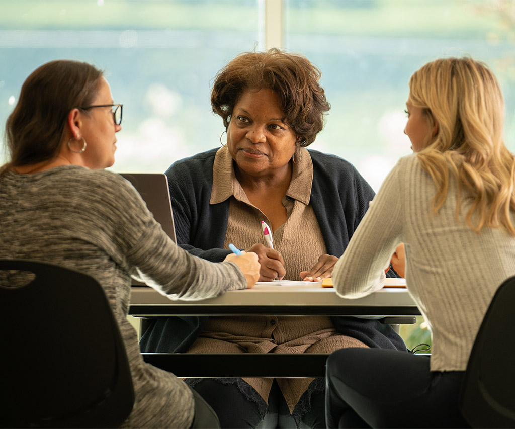 Woman sitting behind a desk, talking to two women on the other side of the desk.