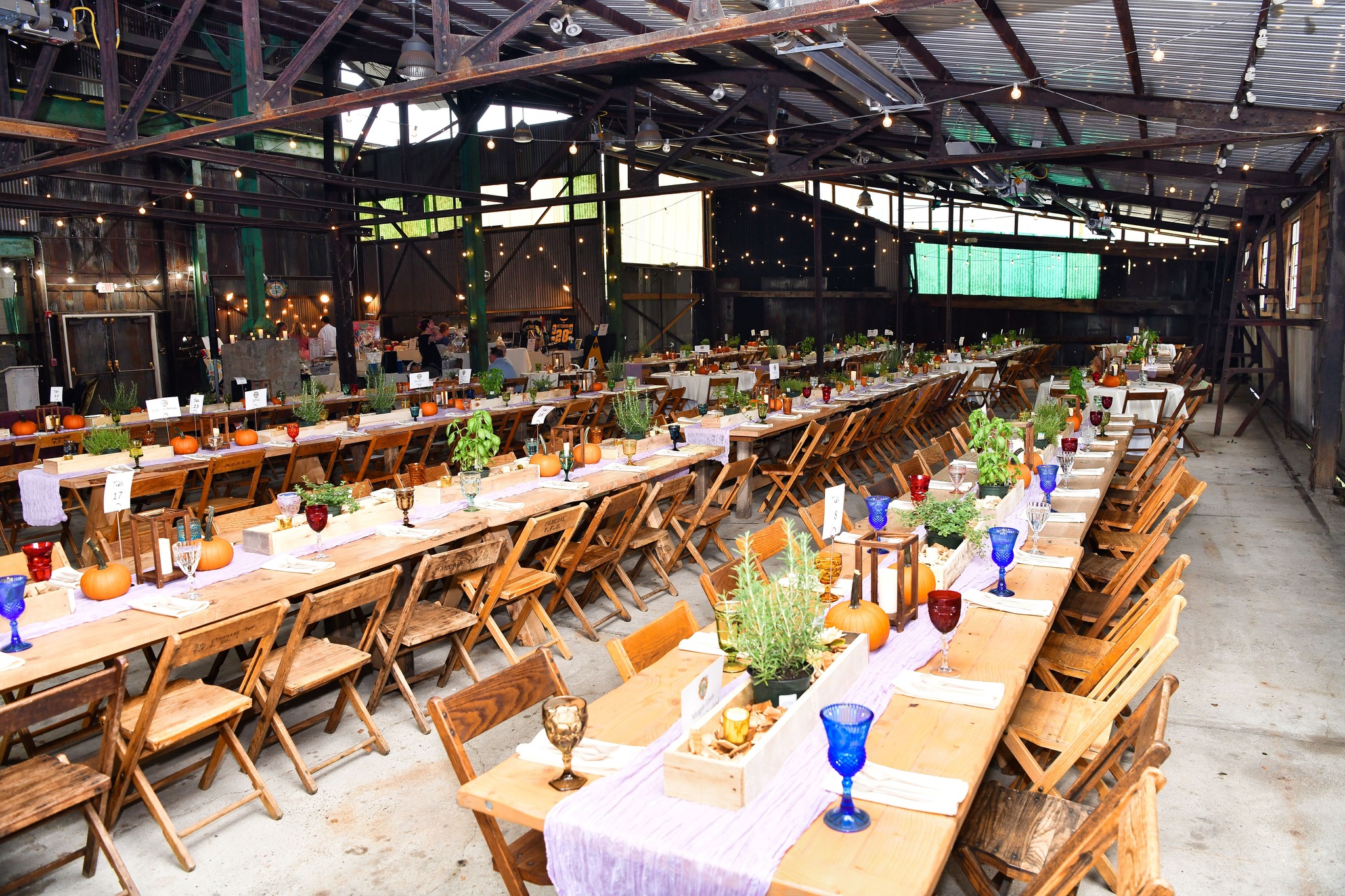 A rustic event venue with long wooden tables decorated with pumpkins, plants, colorful glassware, and string lights hanging overhead.