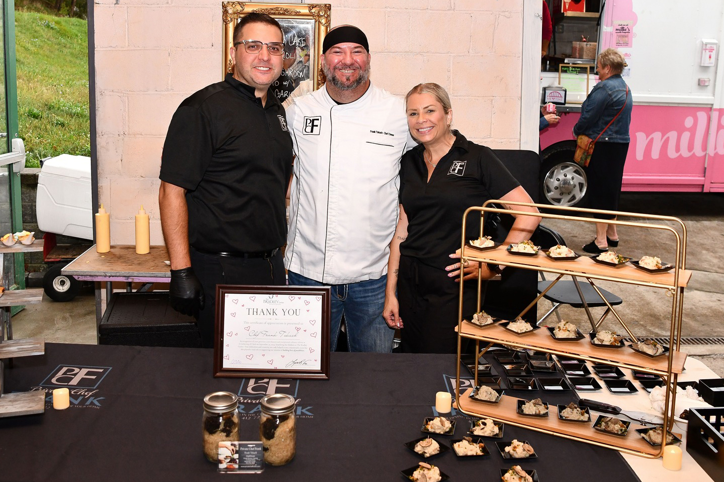 A team of caterers smiles behind a table of beautifully plated appetizers, with a pink ice cream truck visible in the background.