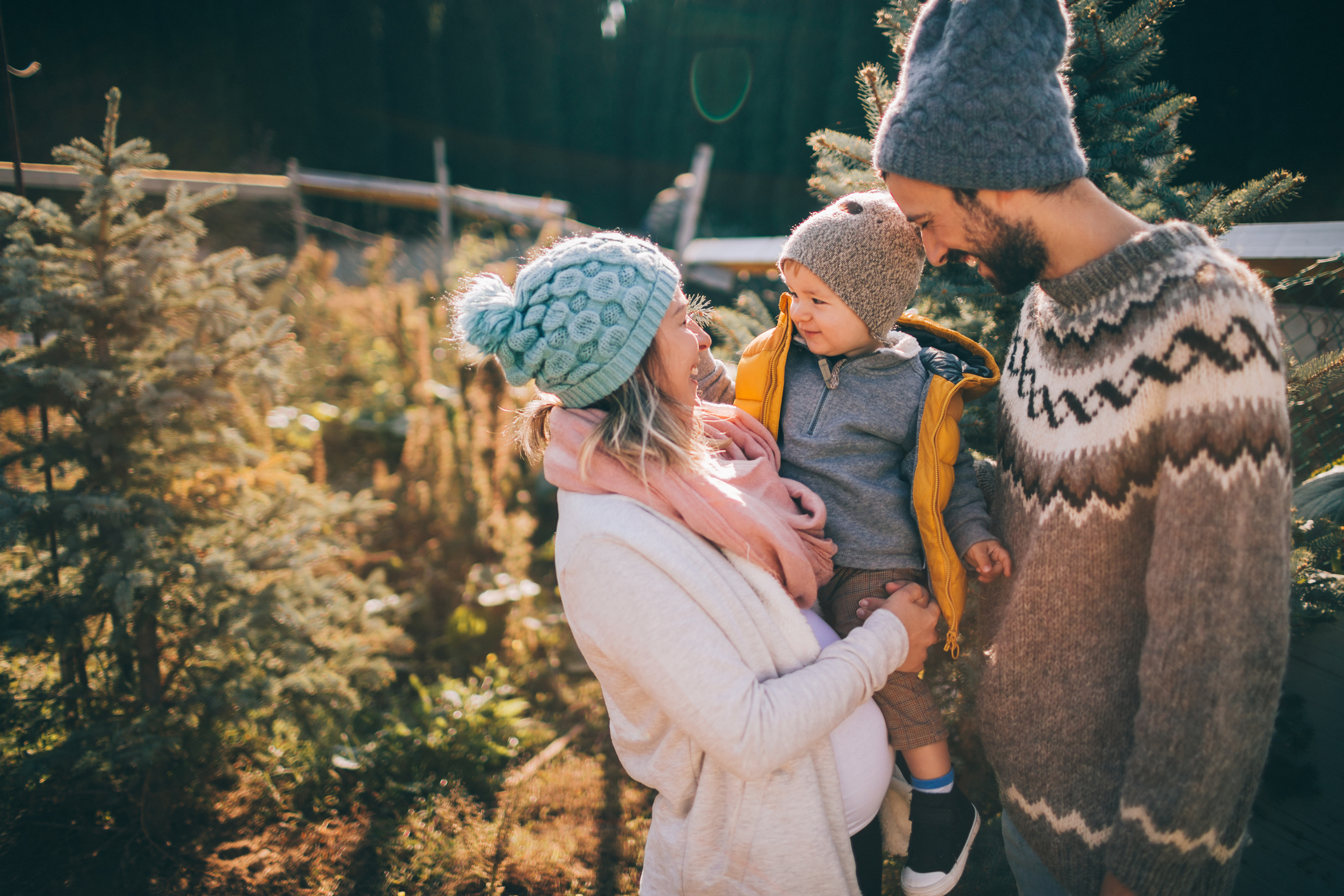 A smiling family poses outdoors with a toddler, wearing warm winter clothing, surrounded by festive evergreen trees.